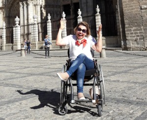 Foto da Laura Martins em frente à Catedral de Toledo, na Espanha. Ela está em uma cadeira de rodas e está com as mãos levantadas. Ela está sorrindo.