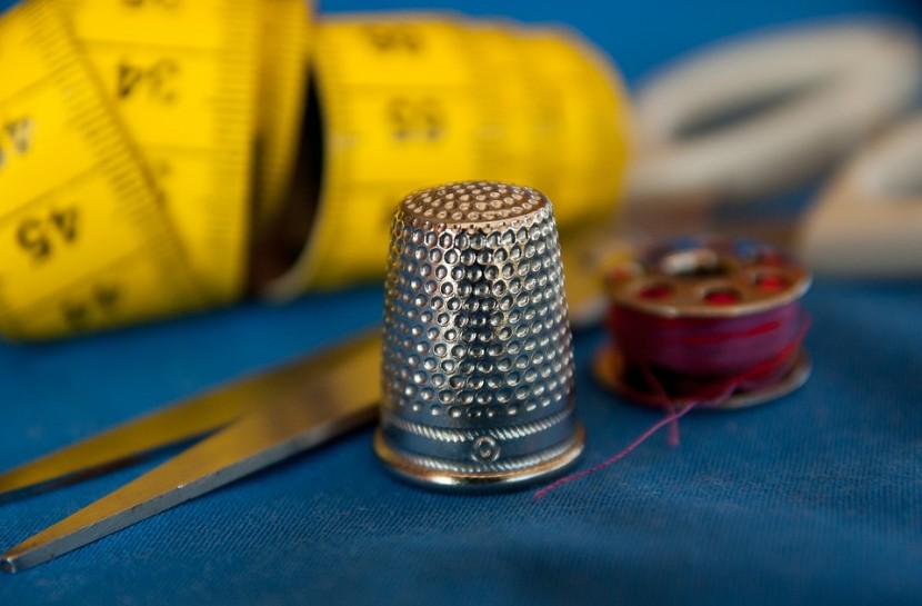 Foto em plano detalhe de um dedal, apoiado em um tecido azul, com uma tesoura, alfinetes e uma fita métrica ao redor