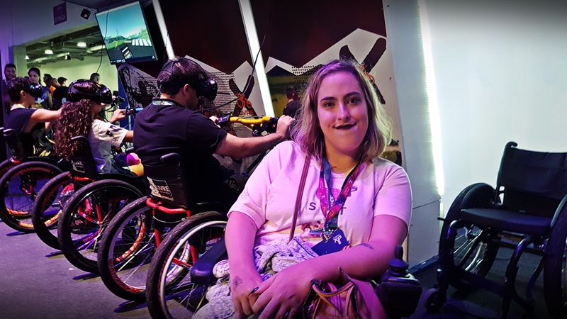 Foto de uma mulher cadeirante na Game XP. Ela olha para a câmera e sorri. Mais atrás, há outros cadeirantes que utilizam óculos de realidade virtual.