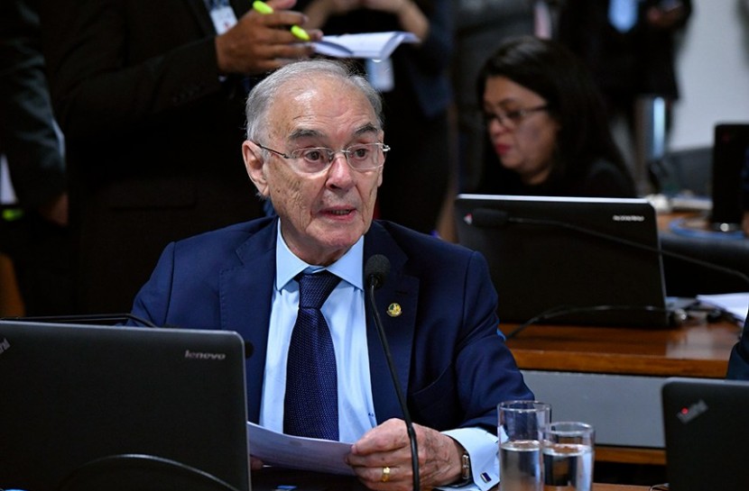 Foto do senador Arolde de Oliveira falando em frente a um microfone no Congresso. Ele é um homem de cabelos brancos, está usando um terno e gravata azuis e uma camisa azul claro.
