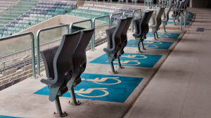 Foto parcial de uma arquibancada de estádio, com poltronas dobráveis e espaços com o símbolo de identificação da pessoa com deficiência, em branco e azul, pintados no chão