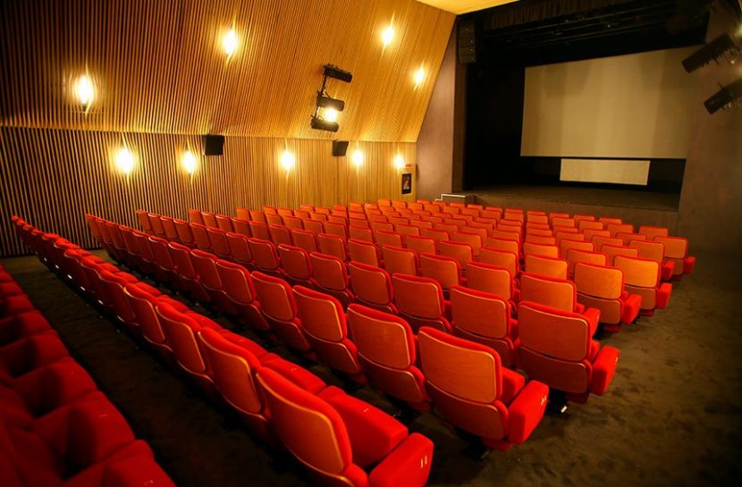 Foto de uma sala de cinema vazia. As cadeiras são vermelhas e há luzes acesas nas paredes.