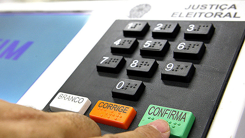 Foto em close do teclado numérico de uma urna eletrônica e a mão de uma pessoa apertando a tecla verde de confirmação.