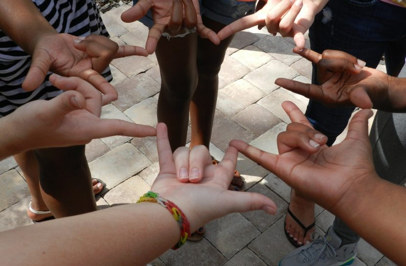 Foto em close de sete mãos diversas fazendo o sinal de amor em Libras, uma ao lado da outra, formando um círculo.