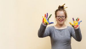 Foto de uma jovem com Síndrome de Down com os braços levantados. As palmas das mãos estão abertas e sujas com tinta. Ela sorri.