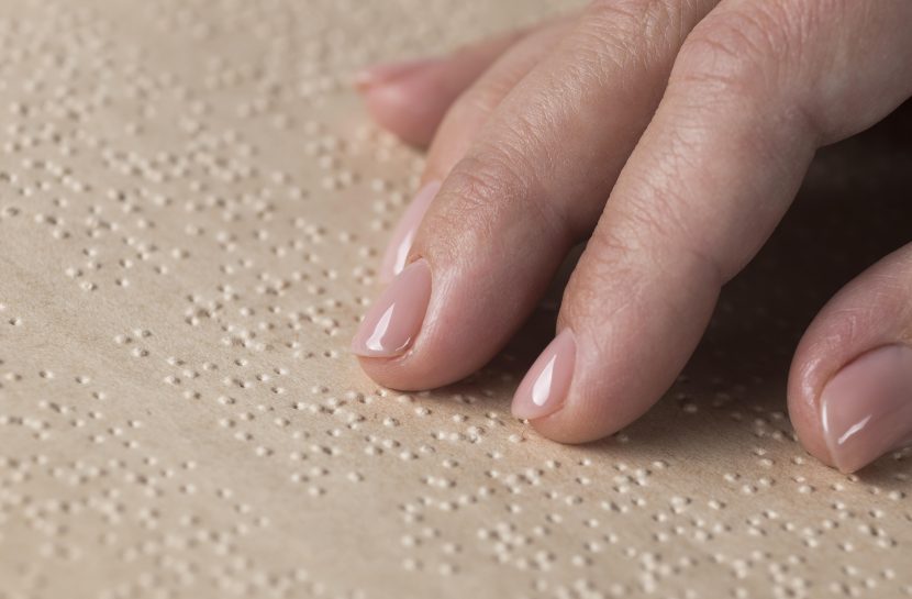 Foto em close dedos sobre papel em relevo de escrita tátil, braille.