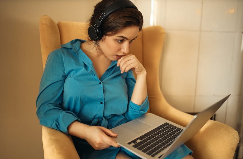 Foto de uma mulher, sentada em uma poltrona, com um laptop em cima das pernas. Ela usa um fone de ouvido e olha para a tela do computador.