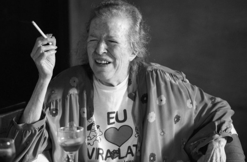 Foto em preto e branco de Hilda Hilst. Ela está com um cigarro em uma das mãos, os olhos fechados e sorrindo.