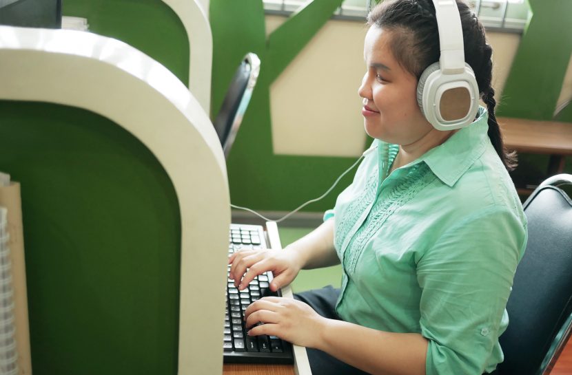 Foto de uma jovem com deficiência visual sando fones de ouvindo e digitando no teclado de um computador.