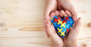 Foto com dois pares de mãos unidos segurando um coração de feltro com estampa de quebra-cabeça nas cores do autismo