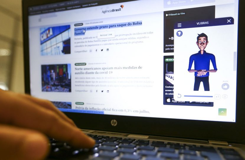Foto da tela de um notebook no site da Agência Brasil, com o avatar da VLibras à direita, no canto da tela. Sobre o teclado do computador, há a mão de uma pessoa, como se estivesse digitando.