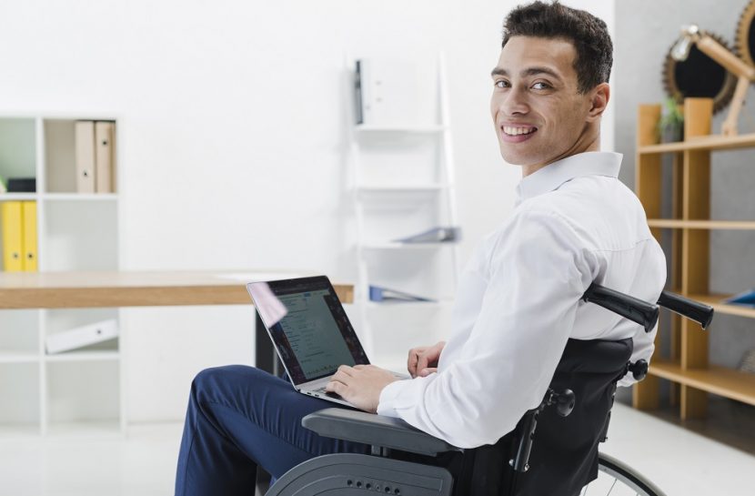 Foto de um homem cadeirante em uma sala de escritório e em seu colo está um laptop. Ele está olhando em direção à câmera.