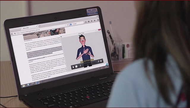 Foto de uma mulher de costas usando um laptop que está em sua frente e sobre uma mesa, nele está aberto um site e em seu lado direito há uma janela com um avatar intérprete de Libras