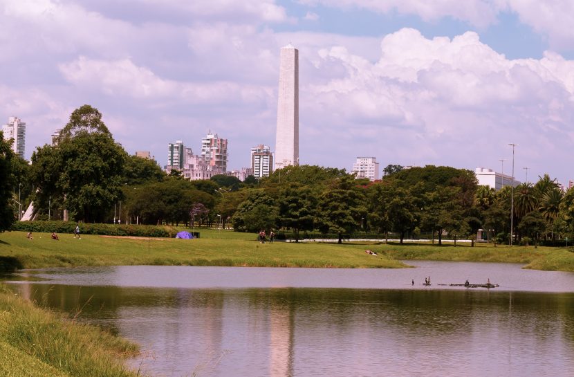 Foto paisagem do Parque do Ibirapuera. O céu está azul e com nuvens brancas, e há muitas árvores, grama e um lago no centro da imagem. Ao fundo, o Obelisco e Monumento Mausoléu ao Soldado Constitucionalista de 32.
