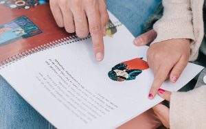 Foto em detalhe de livro infantil em Braille no colo de uma mulher, ao lado dela há uma garota. As duas tocam a página da publicação com a ponta dos dedos