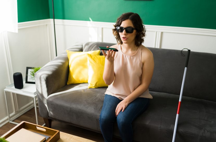 Foto de uma mulher cega sentada no sofá de uma sala com um celular na mão direta, próximo a sua boca. Ao seu lado está uma bengala.