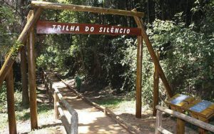 Foto da entrada de uma trilha com paisagem verde ao redor e uma placa de madeira com o texto: “Trilha do Silêncio”.