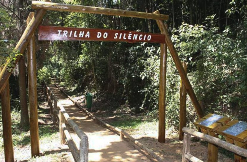 Foto da entrada de uma trilha com paisagem verde ao redor e uma placa de madeira com o texto: “Trilha do Silêncio”.