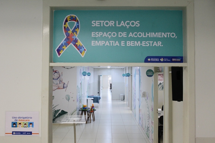 Foto de um corredor do centro de referência em autismo de Uberlândia. Há uma placa na entrada com o texto: “Setor Laços. Espaço de acolhimento, empatia e bem-estar.”, alguns brinquedos sobre uma mesa e diversas portas.