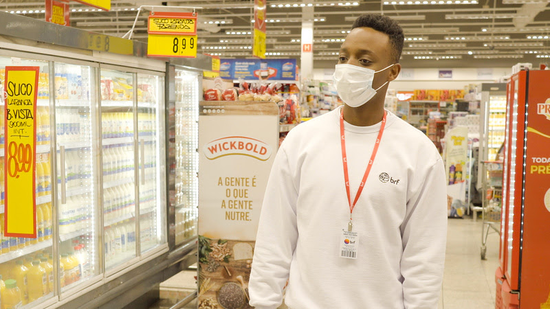 Foto de um jovem em pé em um corredor de supermercado. Ele veste uma blusa branca de manga comprida com o logo da BRF e usa um crachá da empresa.