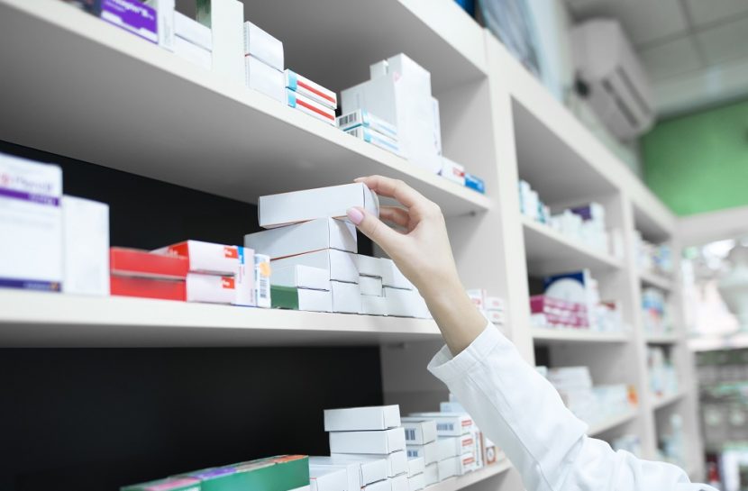 Foto em close da mão de uma farmacêutica pegando uma caixa de remédio de uma prateleira na farmácia.