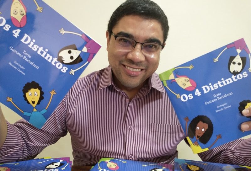 Descrição da imagem: Foto do autor Gustavo Bartolozzi sorrindo e segurando dois exemplares do livro “Os 4 Distintos”. O livro possui uma capa azul, com as quatro personagens principais com as mãos levantadas e sorridentes.