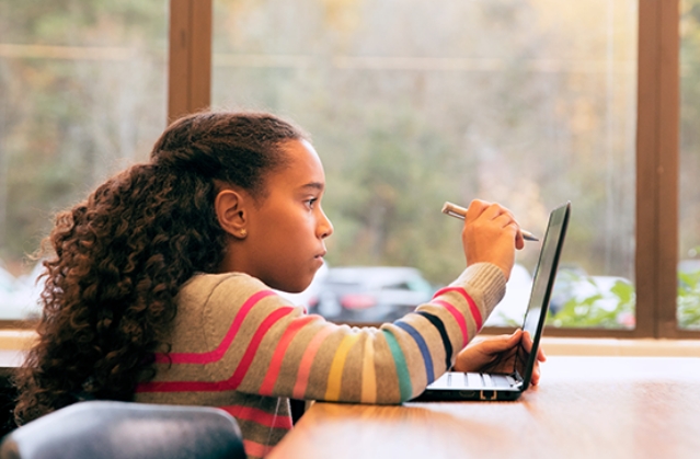 Foto de uma menina de perfil, sentada em uma sala, com uma caneta na mão direita sobre a tela de um laptop.