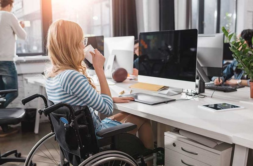 Foto de uma cadeirante em um escritório. Ela está com a mão esquerda em cima do teclado de um computador, enquanto a direita leva uma xícara a boca.