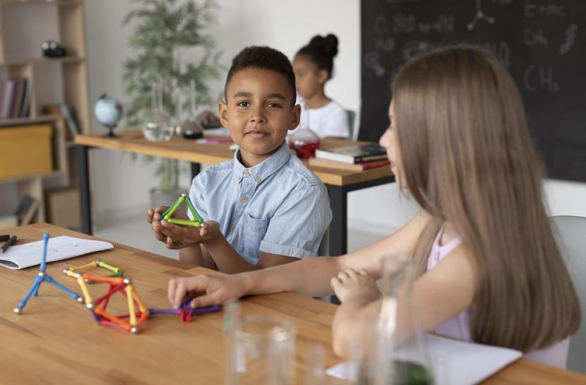 Foto de três crianças em uma sala de aula. Duas delas, um menino e uma menina, estão sentados na mesma mesa e compartilham os instrumentos da aula.