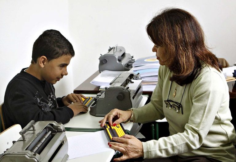 Foto de um menino cego sentando em uma sala com uma professora à sua frente. Ele usa um sorobã, instrumento para fazer cálculos.