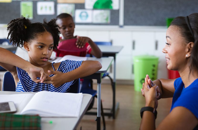 Foto de uma menina e uma mulher em uma sala de aula se comunicando em língua de sinais