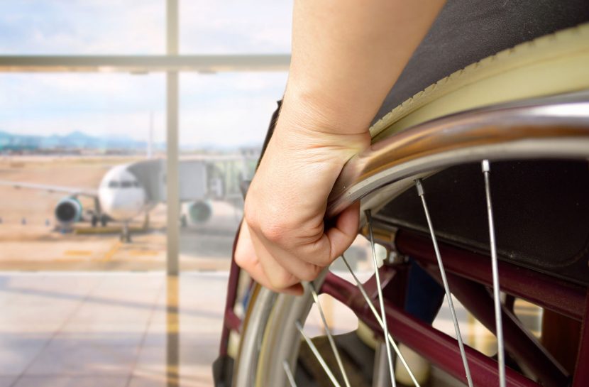 Foto em detalhe de pessoa em uma cadeira de rodas com a mão apoiada na roda. No fundo desfocado, há um avião.