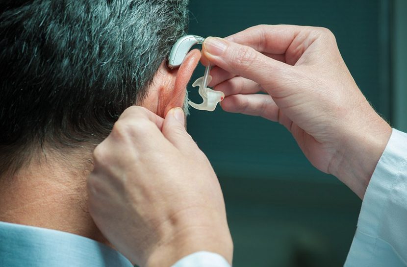 Foto em close de mão colocando um aparelho de surdez na orelha de um homem, que está de costas.